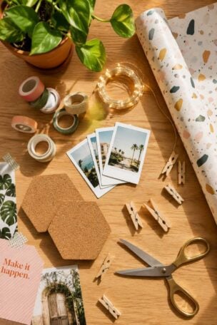 Crafting scene on a dorm room wooden table featuring a potted plant, postcards, washi tape, scissors, string lights, cork coasters, and decorative paper.