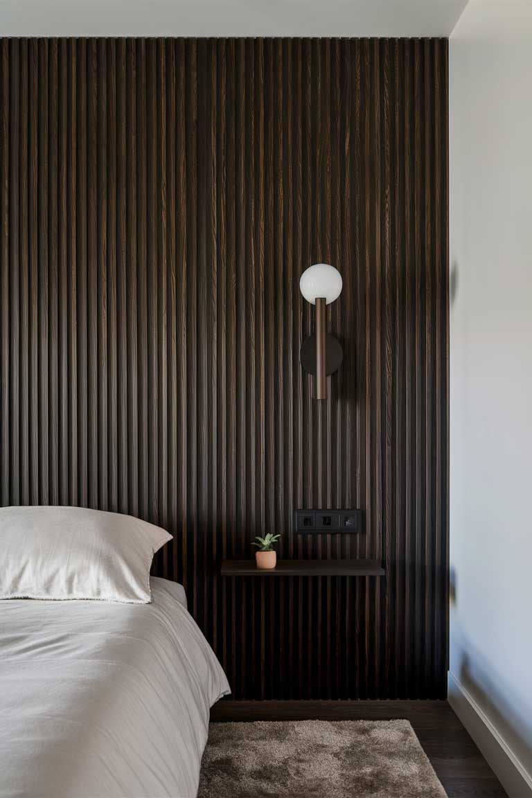 A bedroom wall featuring vertical dark wood slats. The slats extend from floor to ceiling, creating a striking contrast against the adjacent white wall. A minimalist sconce light is mounted on the slatted wall, casting a warm glow.