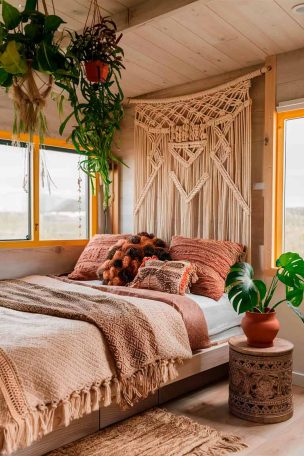 A bohemian-inspired tiny house bedroom with a statement macramé headboard above a low platform bed. The bed is dressed in layers of textured bedding in earthy tones and pattern-rich throw pillows. Hanging plants cascade from the ceiling. A small, intricately carved wooden side table holds a clay pot with a thriving monstera plant.