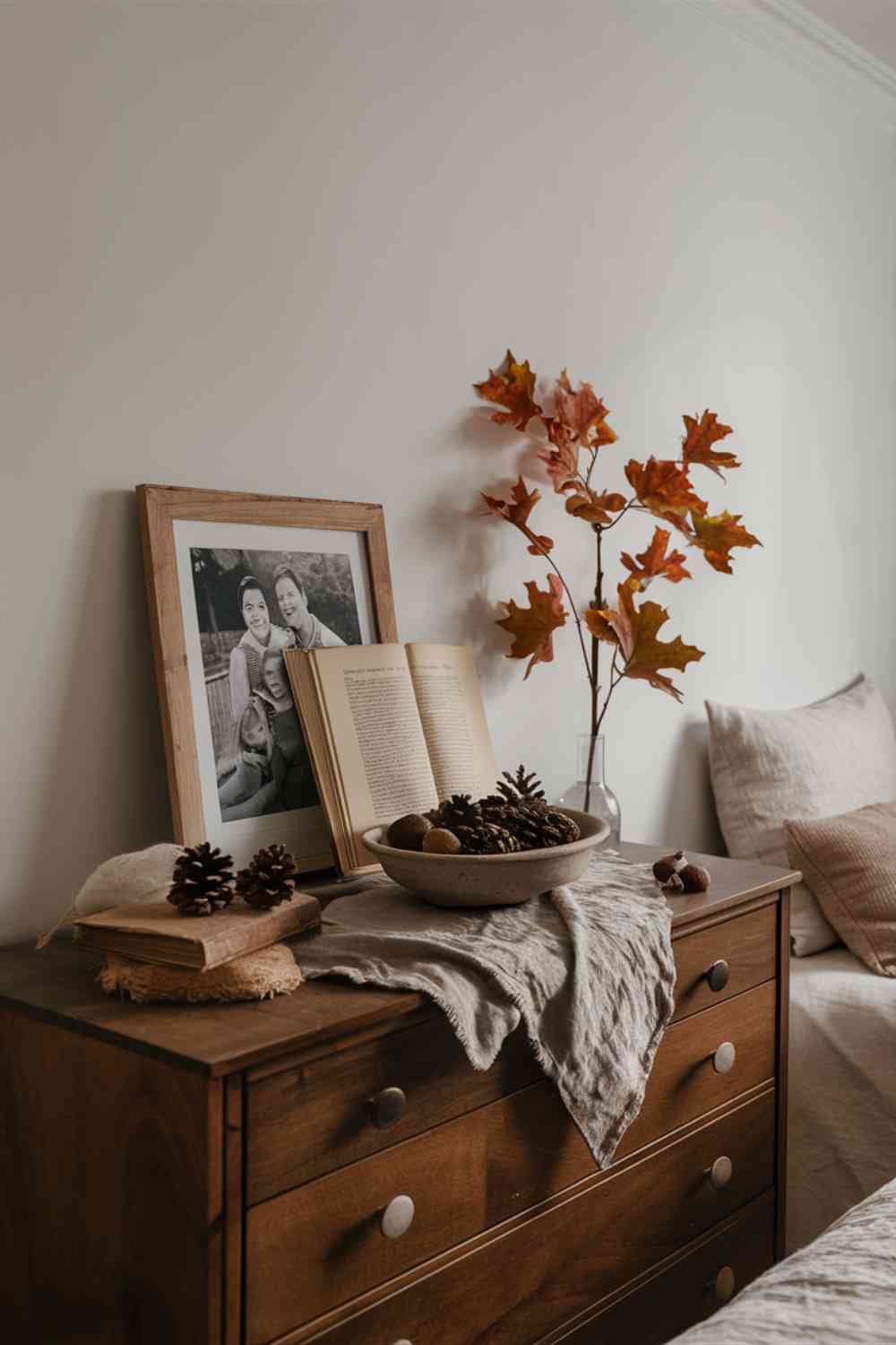 A wooden dresser in a minimalist bedroom. On top of the dresser, there's a carefully curated display of personal items. A simple wooden frame holds a black and white family photograph. Next to it, a vintage book with a warm, leather cover is propped open. A small, hand-thrown ceramic dish holds a couple of pinecones and acorns. The wall above is painted in a soft, warm white, and a branch with autumn leaves in a clear glass vase completes the display. The overall effect is personal and autumnal without being cluttered.