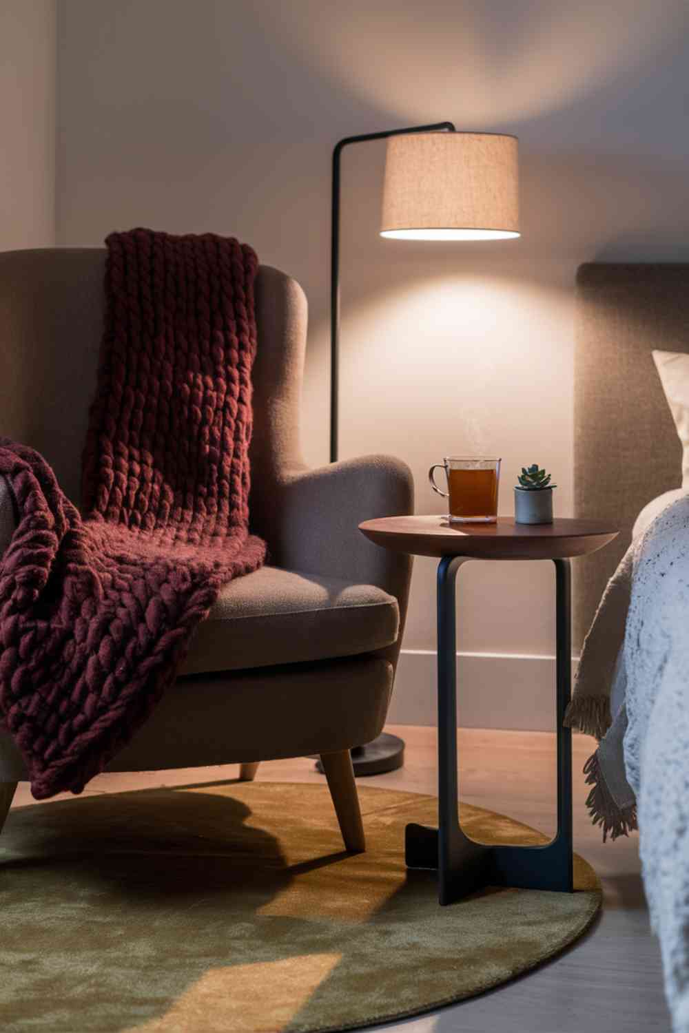 A close-up of a cozy reading nook in a minimalist bedroom. An oversized armchair in a warm taupe color is draped with a chunky knit throw in a rich burgundy shade. A small side table holds a steaming mug of tea, and a soft, circular area rug in a muted mustard tone anchors the space. The lighting is warm and inviting, creating a perfect spot for autumn relaxation.