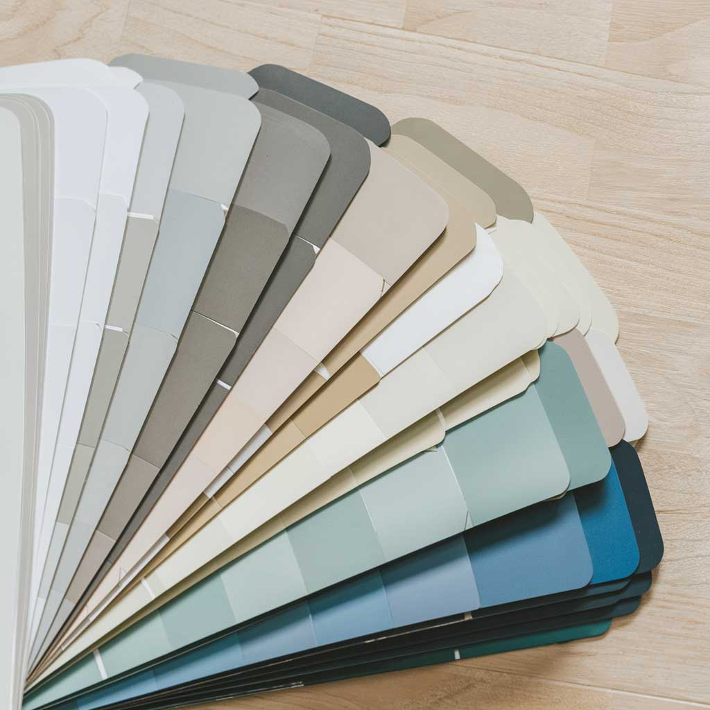 A collection of color swatches arranged in a fan. The colors range from pure white to light gray, soft beige, and pale blue. The swatches are photographed on a light wood surface, reminiscent of Scandinavian flooring.