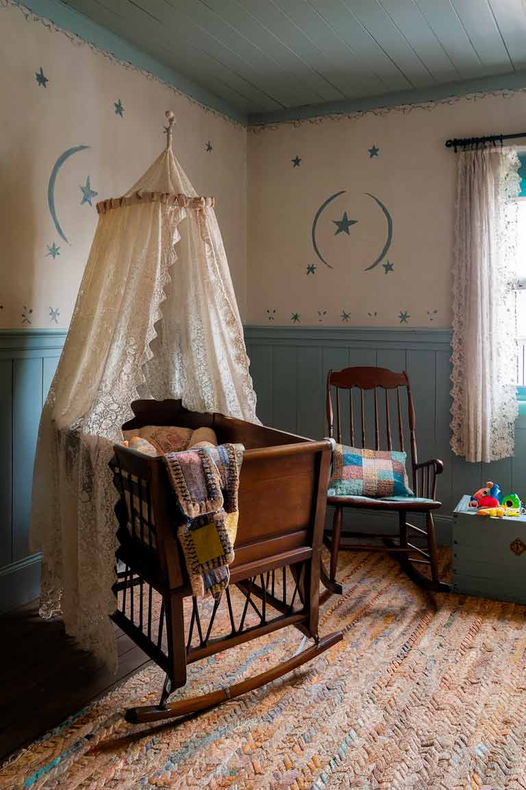 A small colonial nursery with a wooden cradle as the focal point. The cradle is draped with a lace canopy and filled with a soft quilted blanket. A rocking chair sits in the corner, a patchwork cushion on its seat. A small chest painted in a soft blue holds toys and clothes. The walls are adorned with simple stenciled patterns of stars and moons. A braided rug in pastel colors covers the wooden floor, and a window with eyelet curtains lets in soft light.