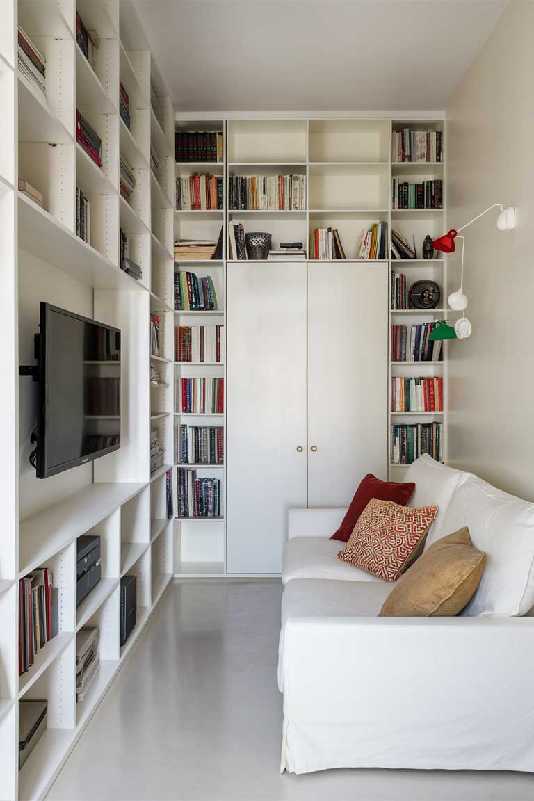 A compact living room with floor-to-ceiling built-in white shelving units flanking a wall-mounted TV. A minimalist white sofa sits opposite, with a few colorful throw pillows adding a pop of color. The room looks organized and spacious despite its small size.