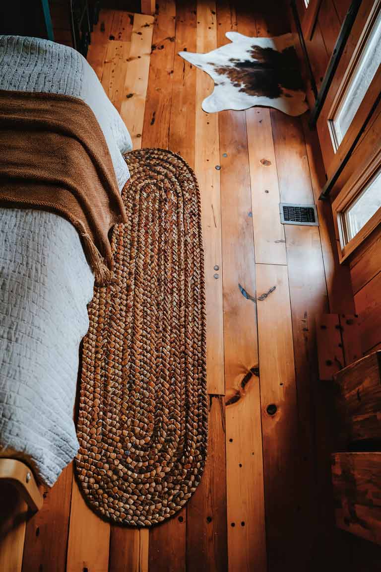 A rustic tiny house bedroom floor featuring wide-plank hardwood in a warm, honey tone. A braided oval rug in earthy colors sits beside the bed, and a small cowhide rug is placed near the door. The contrast of textures adds depth and interest to the space.