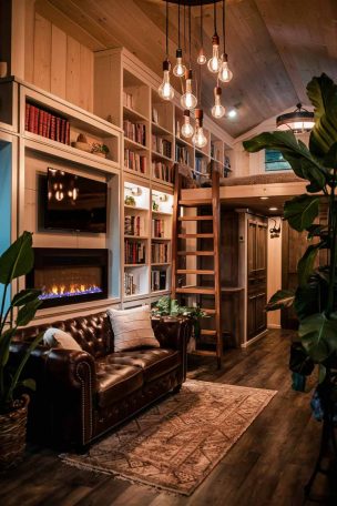 A cozy tiny house living room with a rustic-modern vibe. A small leather loveseat faces a wall-mounted electric fireplace, flanked by built-in bookshelves. A ladder leads to a loft area, visible above. Warm lighting from Edison bulb pendants creates an inviting atmosphere. Large potted plants bring nature indoors.