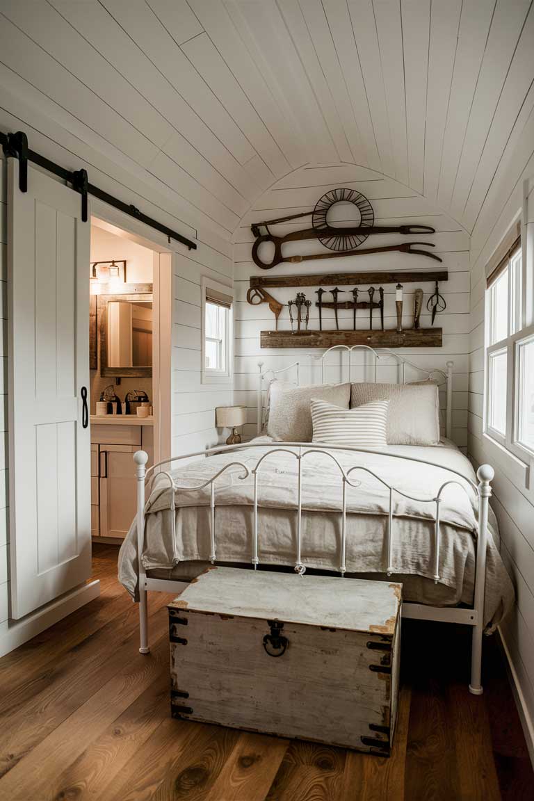 A farmhouse chic tiny house bedroom with white shiplap walls and a sliding barn door. The bed features a white wrought-iron frame dressed in soft linen bedding. Vintage farm tools are artfully arranged on the wall above the bed, and a distressed wooden chest sits at the foot of the bed.