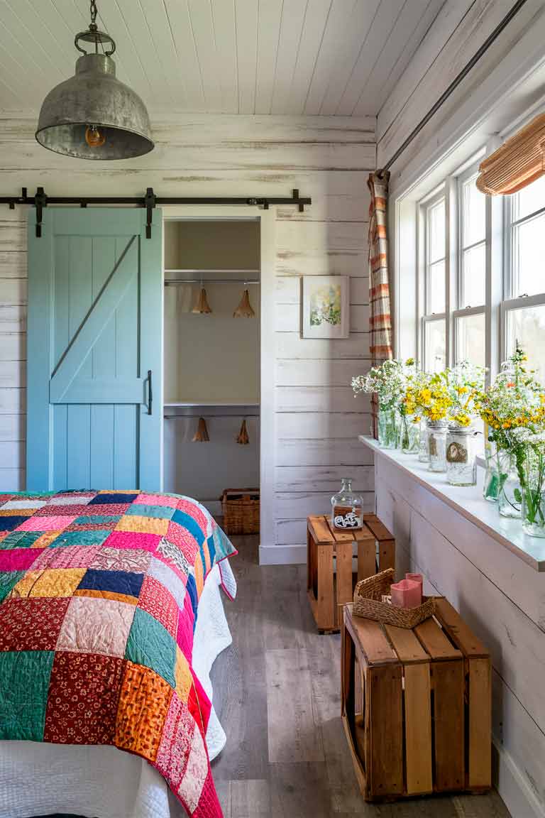 A tiny farmhouse-style bedroom with whitewashed wood walls. A sliding barn door painted in soft blue leads to a small closet. The bed is covered with a colorful patchwork quilt and is flanked by small wooden crates used as nightstands. A galvanized metal pendant light hangs from the ceiling, and mason jars filled with wildflowers sit on a windowsill.