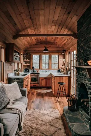 A rustic tiny house interior, showcasing a living area with warm wood tones, a small kitchen visible in the background, and large windows letting in natural light. The space should feel cozy and inviting, with a mix of textures like wood, stone, and soft fabrics.