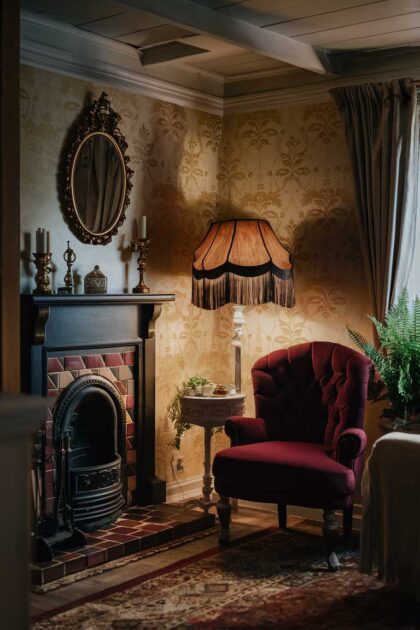 12 Vintage Tiny House Living Room Ideas to Adore 17 A cozy Victorian-inspired tiny house living room corner. The focal point is a small, ornate fireplace with a tiled hearth and a carved wooden mantel. Above the fireplace hangs a gilded oval mirror. A tufted velvet armchair in deep burgundy sits nearby, with a delicate side table holding a vintage lamp with a fringed shade. The wallpaper features a subtle floral pattern in muted gold and cream. A potted fern adds a touch of greenery to the scene.