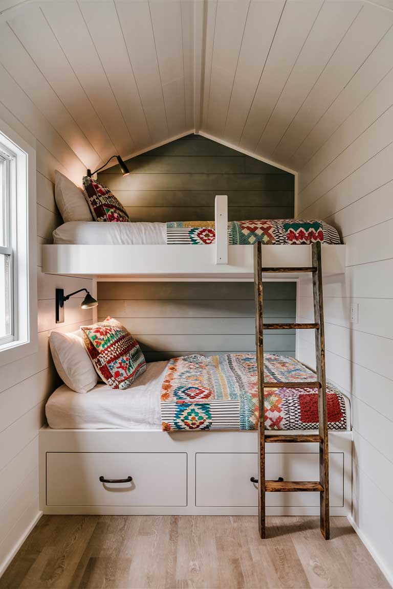 A clever built-in bunk bed design in a tiny house. The bunks are tucked into a wall, with each bed having its own reading light and small shelf. A ladder leads to the top bunk, and the lower bunk has built-in drawers underneath. The beds are dressed in colorful, patterned linens, adding a playful touch to the otherwise neutral room.