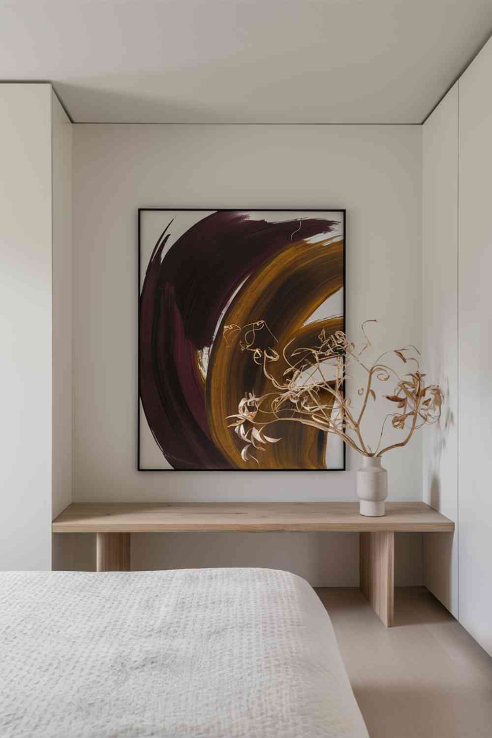 A white wall in a minimalist bedroom featuring a large, simple black frame. Inside the frame is an abstract artwork with sweeping brushstrokes in deep burgundy, mustard yellow, and burnt orange colors reminiscent of autumn leaves. The artwork is the focal point of the wall, with no other decorations around it. Below the artwork, a sleek wooden console table holds a simple white vase with a few branches of dried leaves.