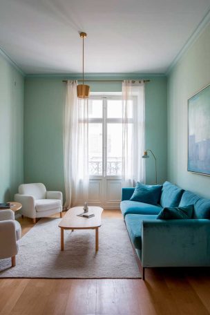 A minimalist living room with soft mint green walls and a sky blue velvet sofa. The room features a light wood coffee table, white accent chairs, and a large window with sheer white curtains. A simple gold pendant light hangs above, and there's a large abstract painting on the wall with shades of blue and green.