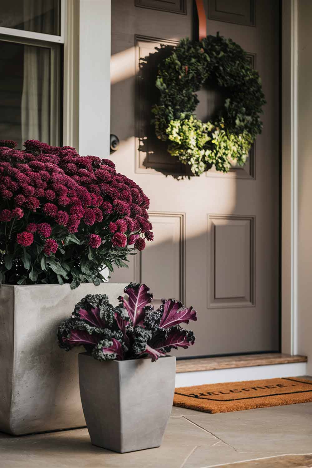 A modern, square concrete planter filled with deep purple mums sits on a porch. Next to it, a smaller planter contains ornamental kale in shades of green and purple. On the door, a simple preserved boxwood wreath hangs from a thin leather strap.