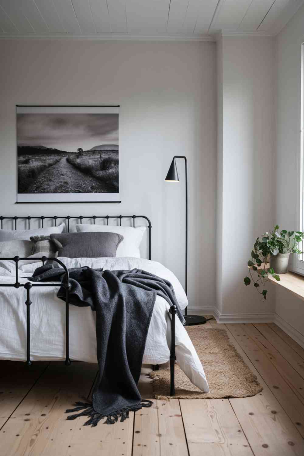 A Scandinavian minimalist bedroom featuring soft white walls and a matte black metal bed frame with delicate, curved details. The bed is dressed in crisp white linens, with a dark gray woolen throw casually draped across the foot, adding texture and warmth. Plush pillows in varying shades of gray and white are layered at the head of the bed. Above the bed, a large black-and-white photograph of a serene Nordic landscape in a thin, simple black frame creates a calming focal point. A sleek black metal floor lamp with a soft, warm glow stands in the corner, its minimalist design mirroring the bed frame. Light, natural oak flooring adds warmth, while a small, woven jute rug under the bed invites coziness. To the side, a floating wooden shelf holds a small potted plant with lush green leaves, a stack of neutral-toned books, and a minimalist ceramic vase, introducing a touch of nature and subtle color to the space. The overall atmosphere is serene, warm, and inviting."