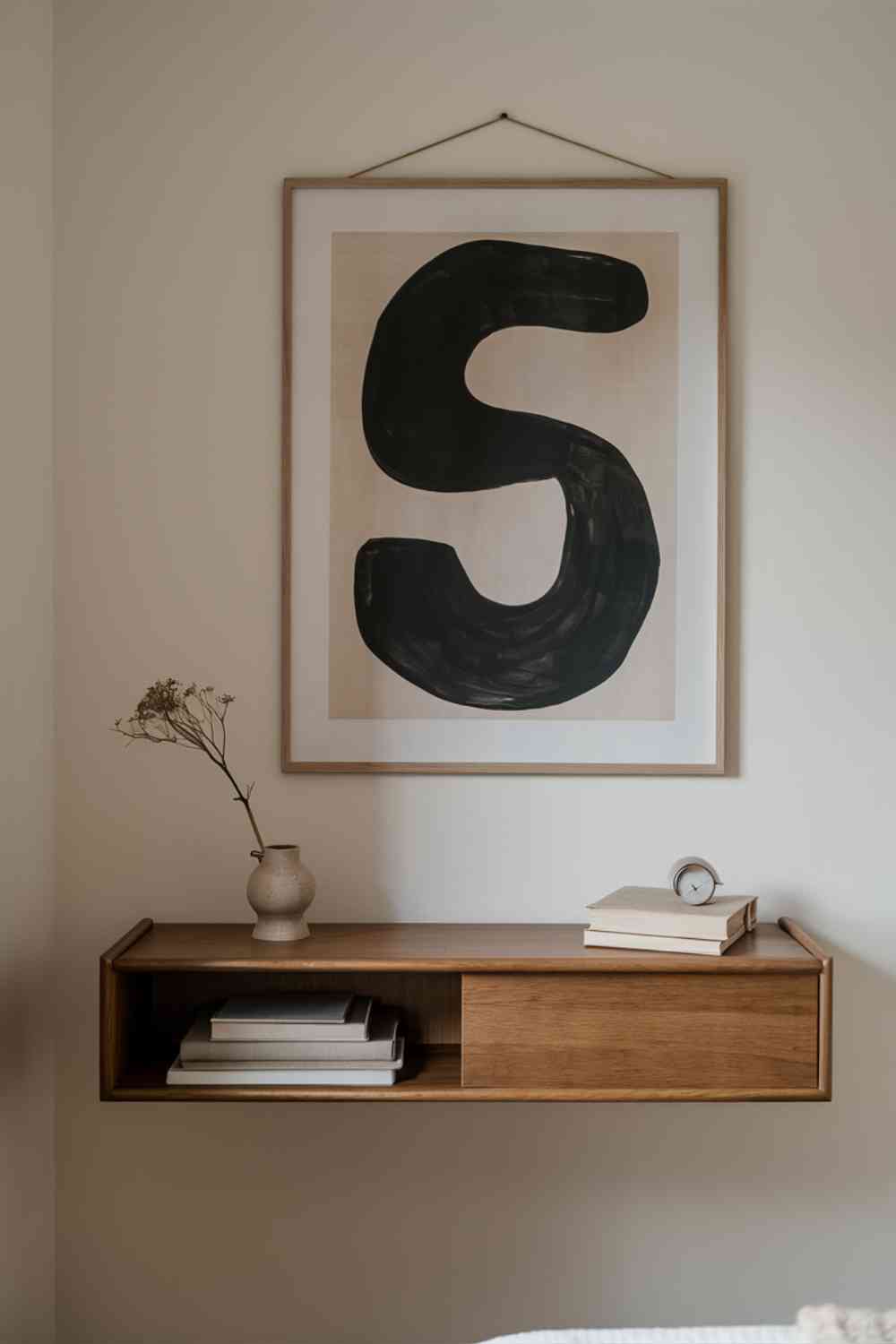 A Scandinavian minimalist bedroom wall featuring a large, abstract black and white print in a simple frame. Below it, a wooden floating shelf holds a few carefully curated items: a small ceramic vase with a single stem, a minimalist clock, and a small stack of books with neutral-colored spines.