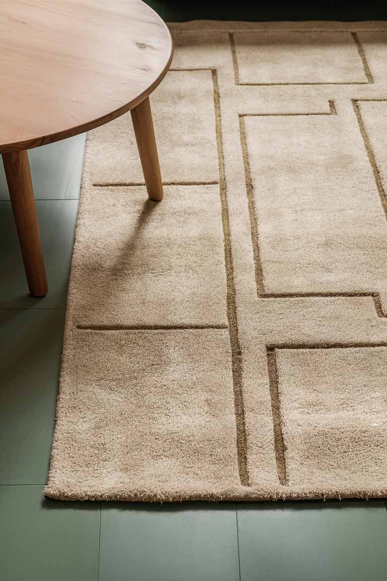 A section of a sage green living room floor showing the edge of a large area rug with a subtle, minimalist geometric pattern in neutral tones.