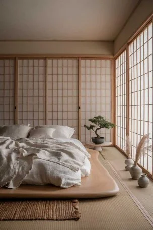 A wide-angle view of a serene Japandi bedroom with a low wooden platform bed, white linen bedding, and shoji screens filtering soft natural light. The room features a neutral color palette with accents of light wood and a single potted bonsai tree on a minimalist side table.