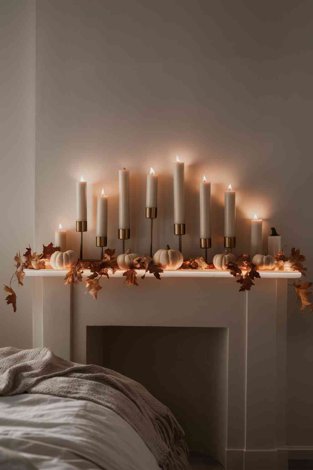 A minimalist fireplace mantel in a bedroom, painted white. On the mantel, there's an asymmetrical arrangement of pillar candles in varying heights, all in warm ivory tones. The candles are placed in simple, modern brass holders of different sizes. A few small white pumpkins are interspersed among the candles. The warm glow of the lit candles creates a cozy, intimate atmosphere in the room.