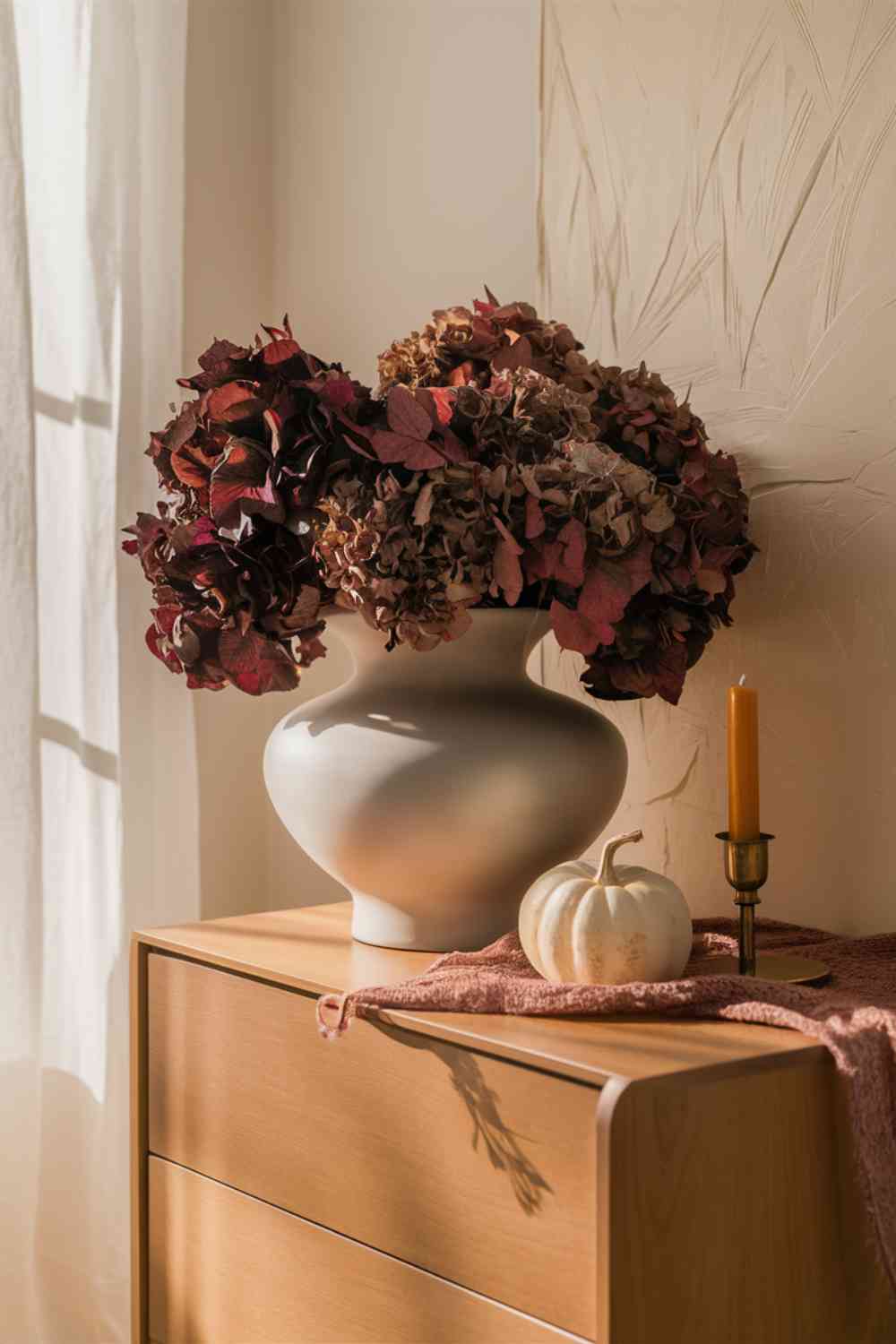 A sleek, white ceramic vase on a minimalist wooden dresser, filled with an arrangement of dried hydrangeas in muted shades of burgundy and deep purple. Next to the vase, a small white pumpkin sits alongside a brass candle holder with a warm, glowing candle. The wall behind is a soft cream color, and warm, diffused light creates a cozy autumn atmosphere.