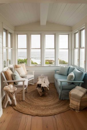 A small, airy living room with large windows overlooking the ocean, featuring soft white walls, natural wood flooring, and furniture in shades of sandy beige and soft blue. The room is decorated with coastal accents like seashells, a driftwood coffee table, and a large, woven jute rug.