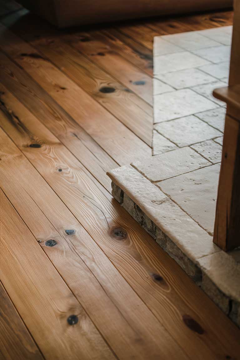 A close-up view of rustic flooring in a tiny living room. The image shows wide wooden planks with a distressed finish, their warm tones, and natural grain patterns visible. In one corner, the wood transitions to stone tiles, showcasing how these two materials can be combined in a rustic interior.