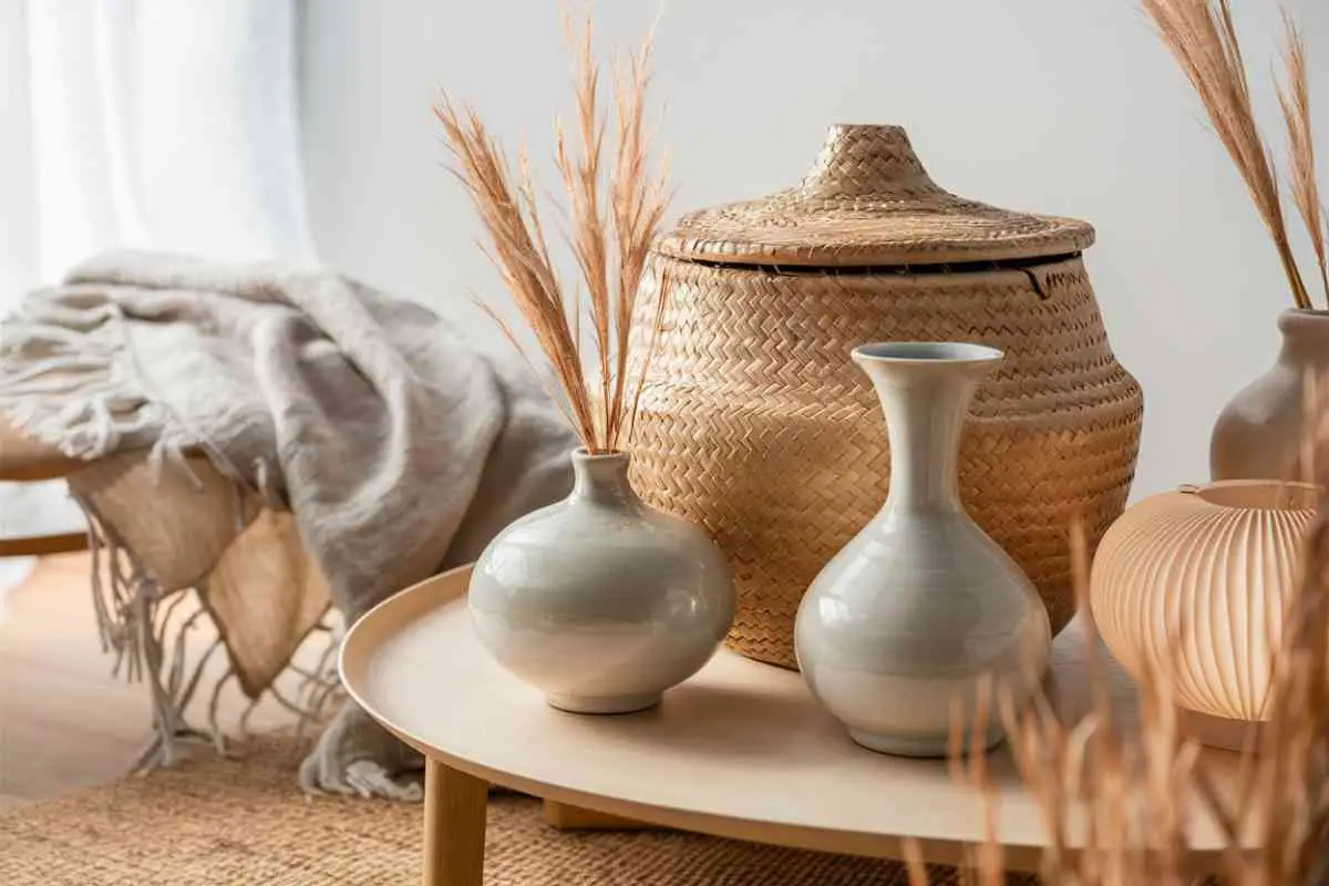 A close-up shot of a stylish Japandi-inspired arrangement. The scene includes a natural woven basket with a lid, positioned next to a pair of elegant, handcrafted ceramic vases with soft, muted glazes. One vase is tall and slender, while the other is shorter and round. The background features a gently textured, light-colored linen throw draped across a low wooden table. On the table, there is also a paper lantern with a delicate, subtle pattern. The overall composition emphasizes the texture and natural materials, with a clean, minimalist backdrop to keep the focus on the items. The lighting is soft and diffused, highlighting the natural colors and textures of each piece.