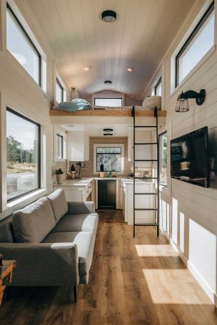 A sunlit modern tiny house living room with a loft bedroom above. The space features a compact sofa, wall-mounted TV, and a ladder leading to the loft. Large windows and light colors create an airy feel.