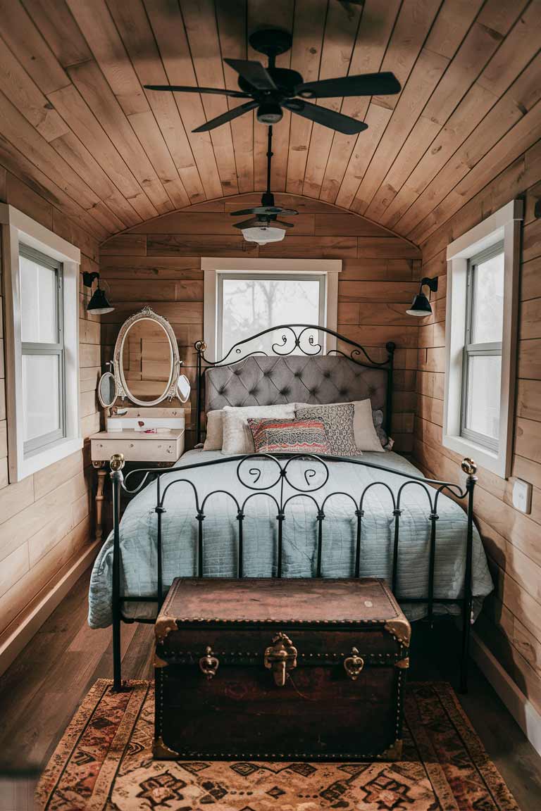 A tiny house bedroom with a wrought iron bed frame and tufted headboard. A small vintage vanity with a mirror is tucked into one corner, and an antique trunk sits at the foot of the bed.