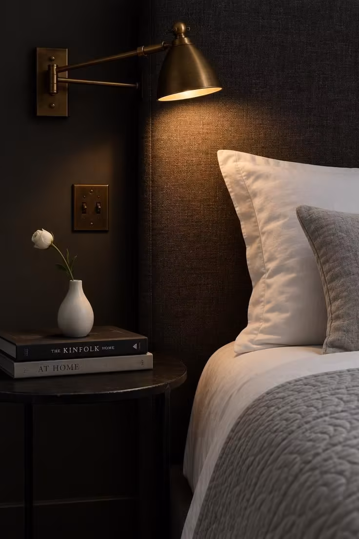 13 Calm Adult Bedroom Ideas for Women to Feel at Home 4 Brass swing-arm sconce beside a charcoal upholstered headboard, blackened steel side table with books and a bud vase, and crisp white bedding in low warm light.