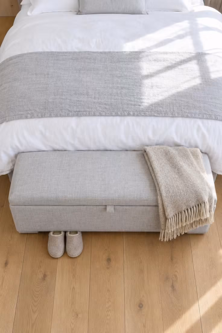 13 Calm Adult Bedroom Ideas for Women to Feel at Home 8 Upholstered pale grey storage bench at the foot of a bed with a folded wool throw and slippers underneath in bright midday light.