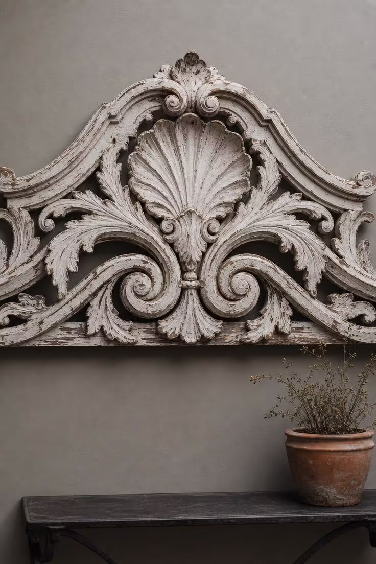 Victorian carved wood pediment with chippy white paint and acanthus leaf motifs mounted flat on a warm grey wall under soft skylight diffusion