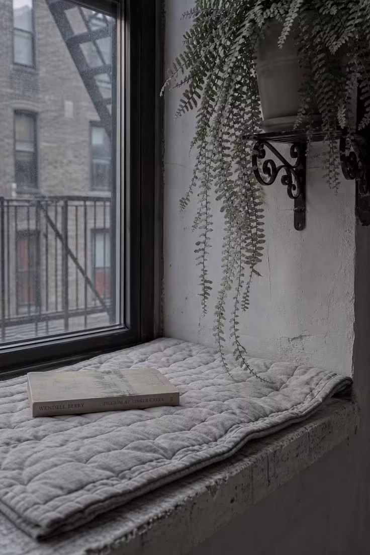 13 Cozy Apartment Reading Nook Ideas for Small Spaces 9 Quilted dove seat pad on a stone window ledge beside a fire escape with trailing fern and cool overcast light