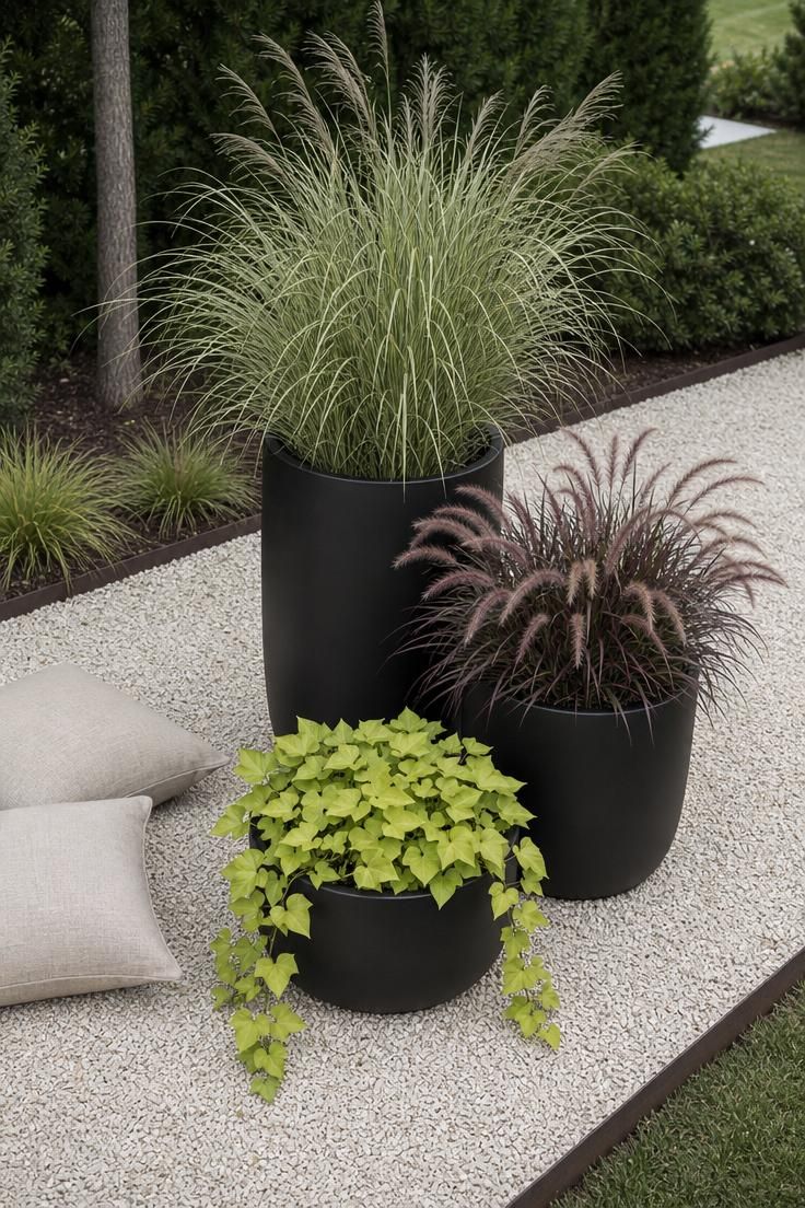 Three matte black fiberglass planters at staggered heights on a crushed limestone gravel patio with ornamental grasses in soft diffused light.