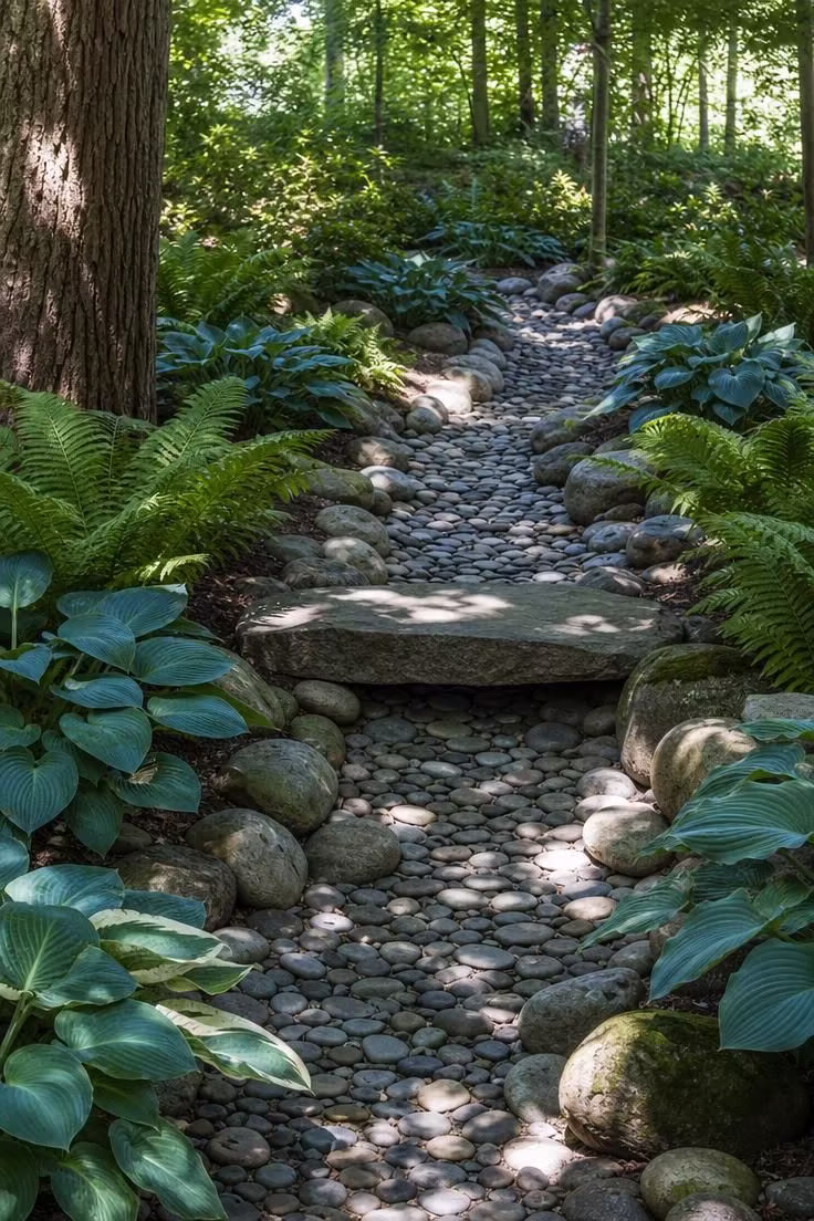 13 Clever Backyard Ideas on a Budget That Feel Polished 12 River rock dry creek bed winding through blue-green hostas and fern fronds in bright midday dappled light