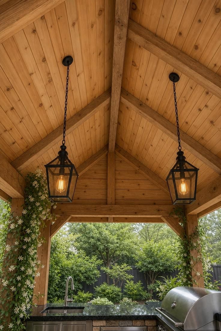 Cedar plank ceiling inside a gabled roof pavilion over a backyard cooking station with pendant lights and jasmine on a post
