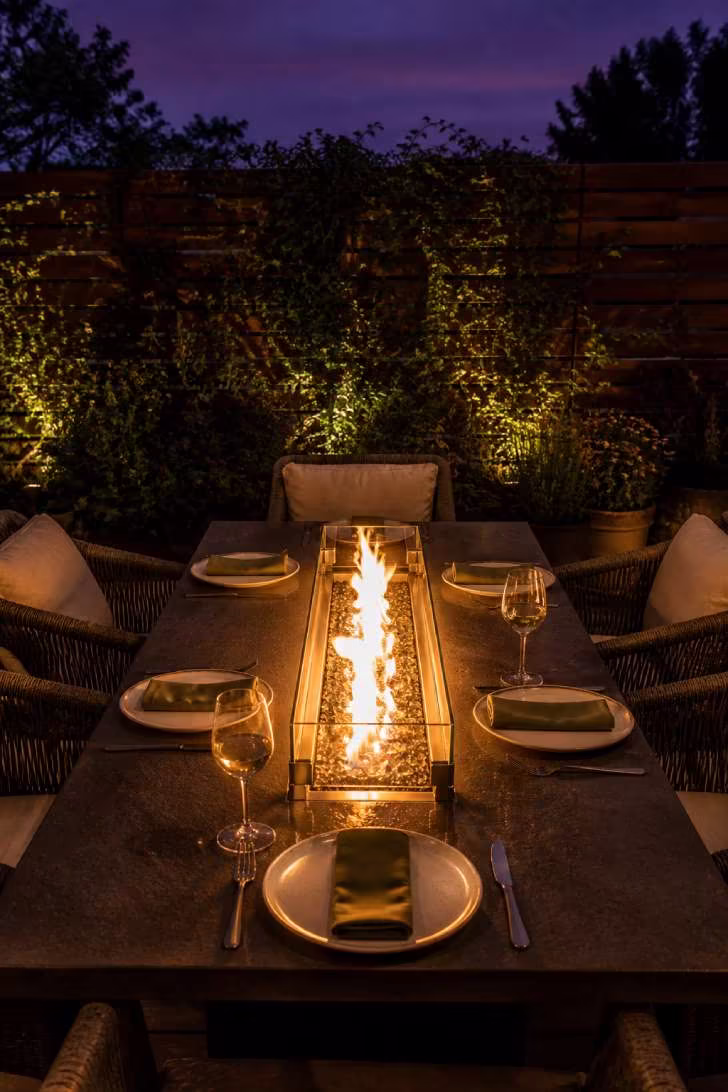 A dark concrete rectangular fire table set for dinner with white stoneware, olive linen napkins and wine glasses, surrounded by woven chairs under twilight sky