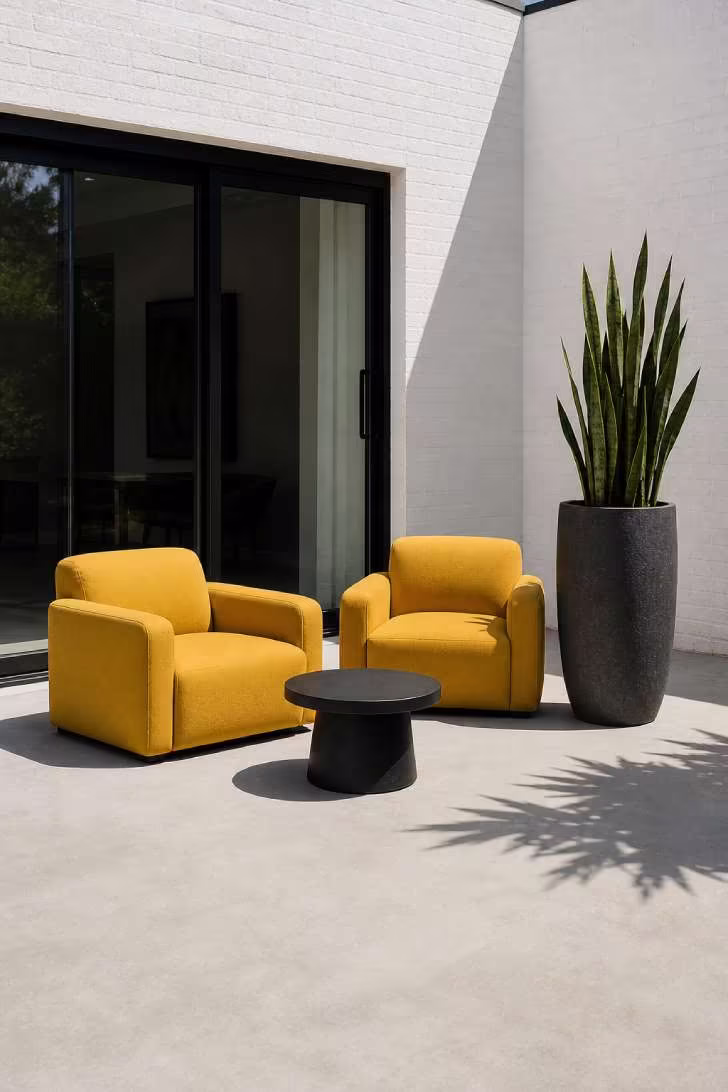 A plain gray concrete patio with two mustard yellow lounge chairs, a matte black side table, and a single tall charcoal planter holding a snake plant in direct midday sun
