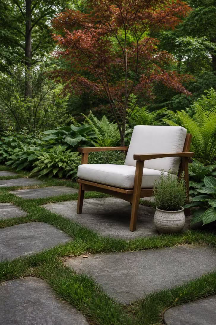 Large bluestone slabs set into a green lawn with grass growing between them, a mid-century acacia lounge chair with ivory cushion, and a lush garden of hostas and ferns in soft overcast light