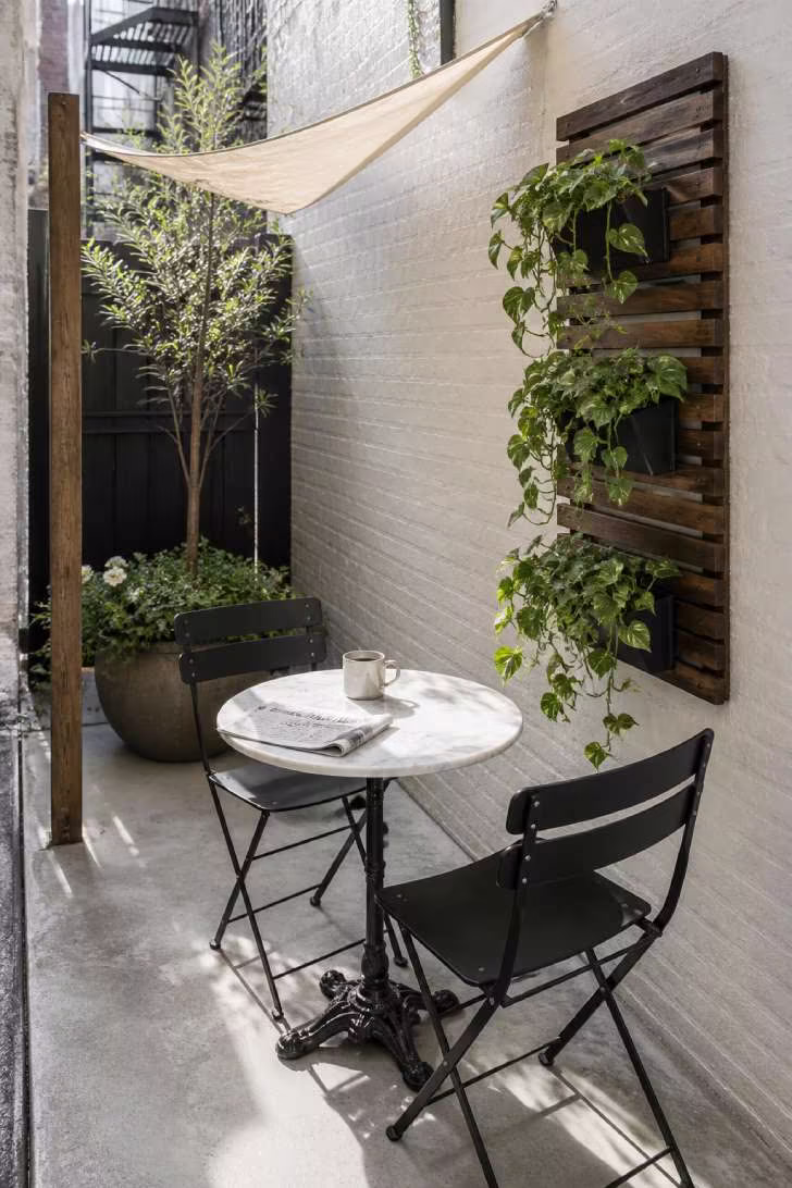 A small corner patio with a white marble bistro table, two black folding chairs, a vertical wall planter, and a linen shade sail in bright morning light