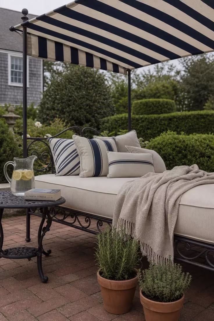 11 Cozy Backyard Reading Nook Ideas for Quiet Escapes 4 Wrought iron daybed with navy and cream striped canvas canopy on herringbone brick pavers with terracotta herb pots and overcast lighting.