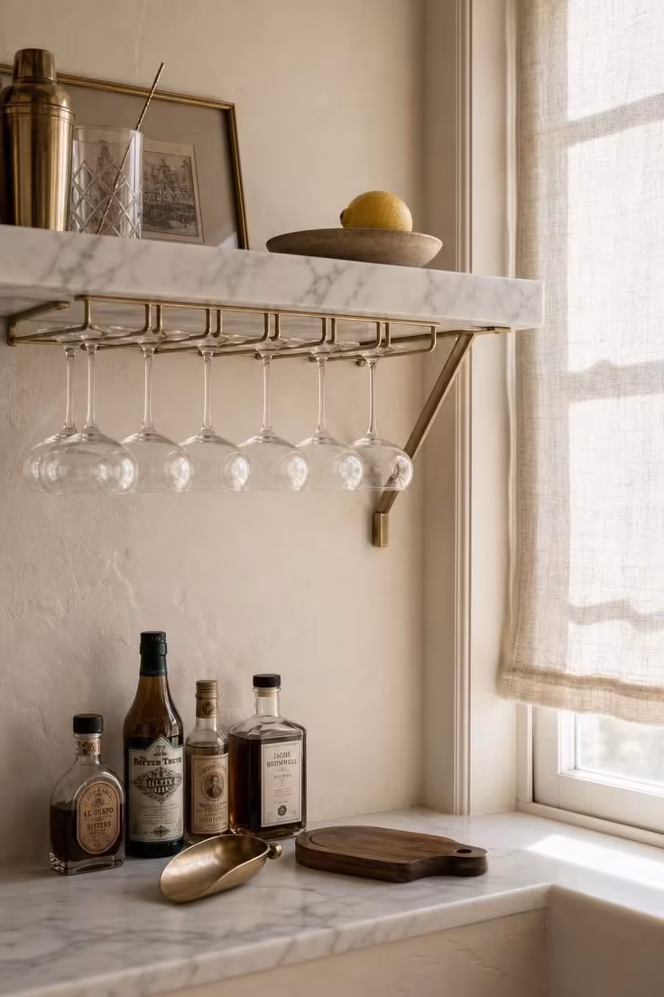 11 Cozy Bar Ideas for Home Living Rooms You'll Love 5 Marble shelf with brass stemware rack hanging coupe glasses above a deep window sill with bitters bottles