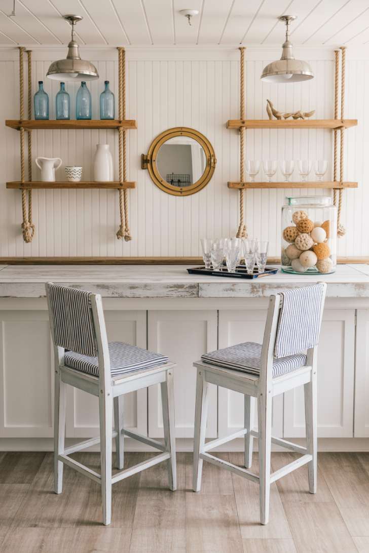 A nautical-themed basement bar with whitewashed wood counter, rope-hung shelves, porthole mirror, and navy ticking-stripe stools