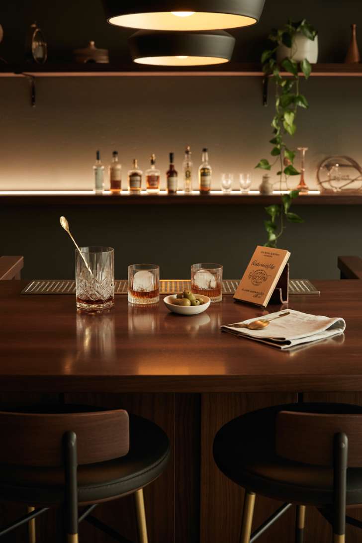 An overhead view of a dark walnut basement bar with crystal mixing glass, whiskey on the rocks, leather-bound recipe book, and warm pendant lighting against a charcoal wall