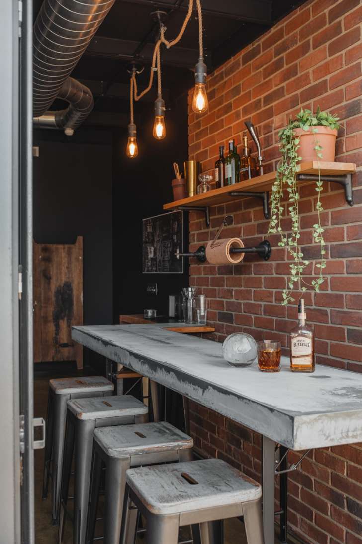 An industrial basement bar with a poured concrete counter, exposed brick wall, black pipe shelving, and Edison bulb pendants at staggered heights