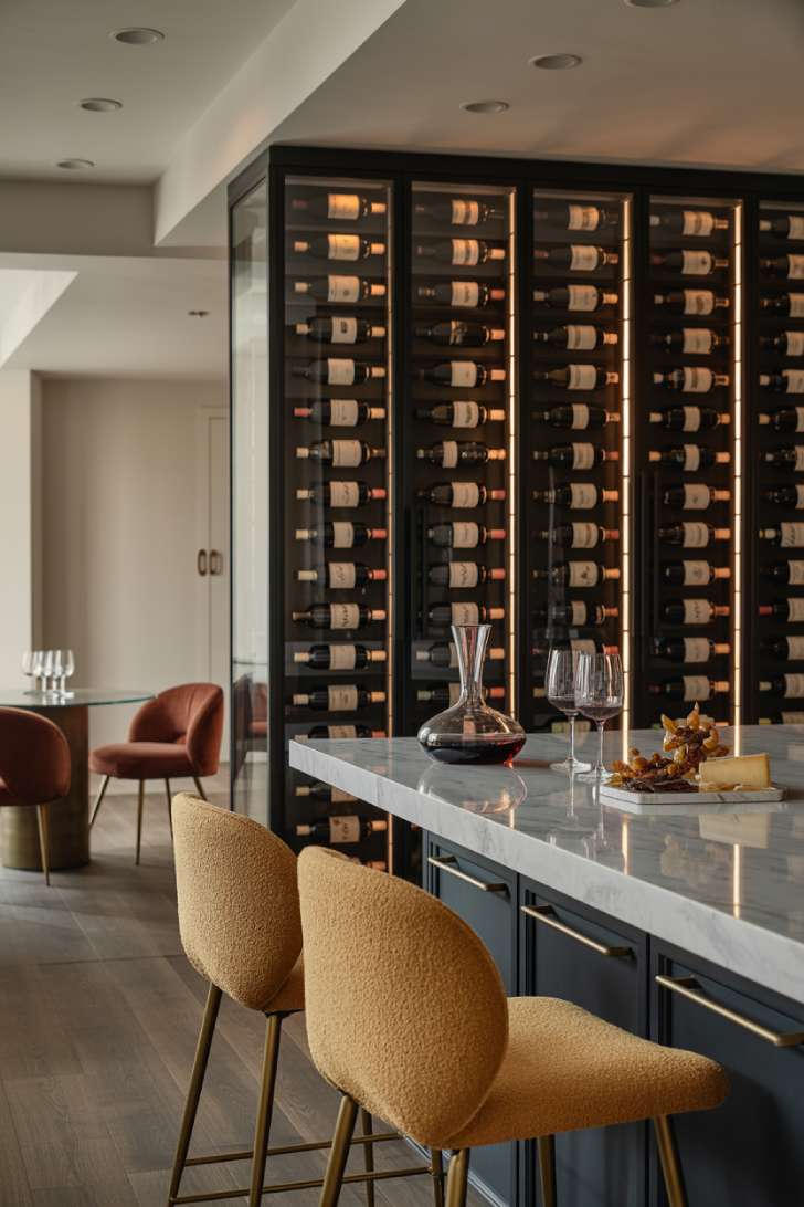 An elegant wine bar with a glass-enclosed backlit wine wall, honed marble counter, navy cabinetry, and plush boucle bar chairs