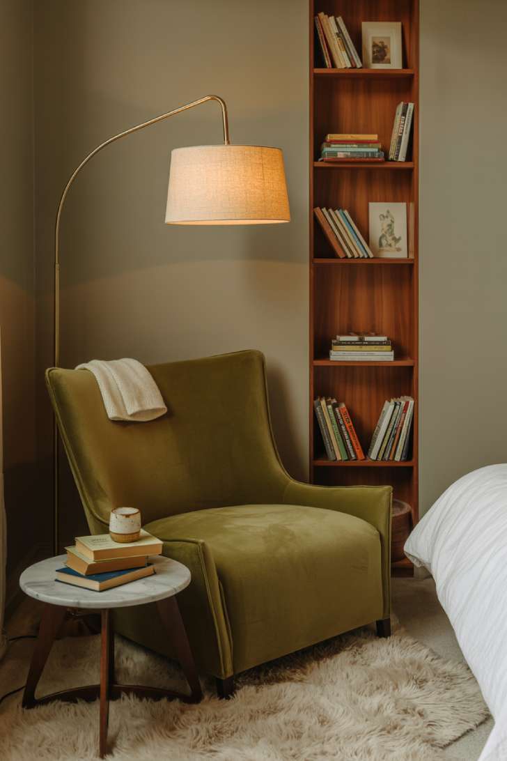Cozy reading nook corner in a basement bedroom with olive green velvet armchair, brass floor lamp, and wall-mounted walnut bookshelf