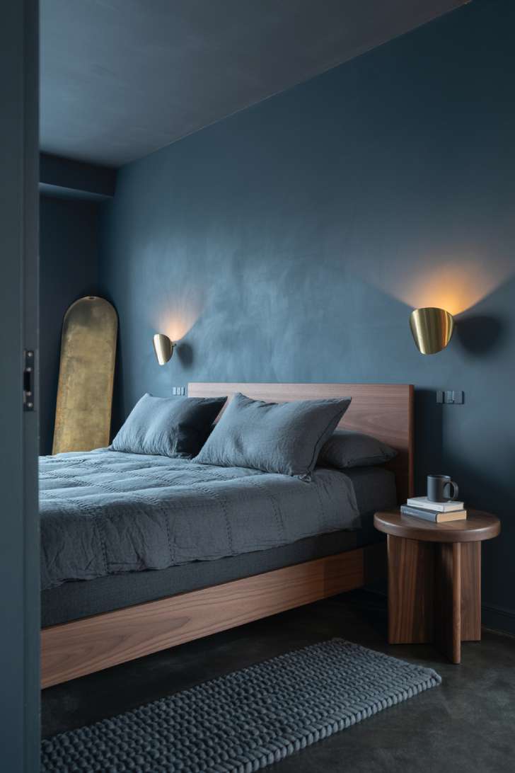 Dark charcoal-blue basement bedroom with brass sconces, low walnut bed frame, and moody atmospheric lighting