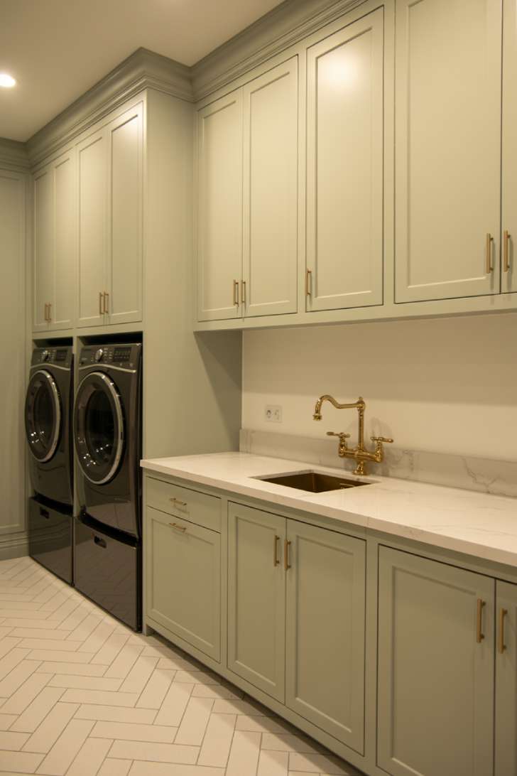 Floor-to-ceiling sage green cabinets with brass hardware concealing washer and dryer in a finished basement laundry room