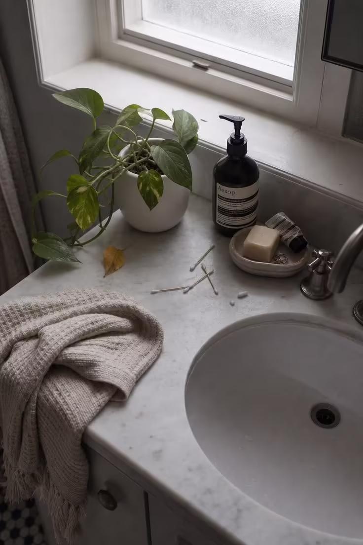 7 Mistakes That Make a Bathroom With Plants Look Messy Instead of Calm 1 Overhead view of a bathroom vanity with a small pothos plant beside scattered toiletries and a drooping leaf in cool morning light